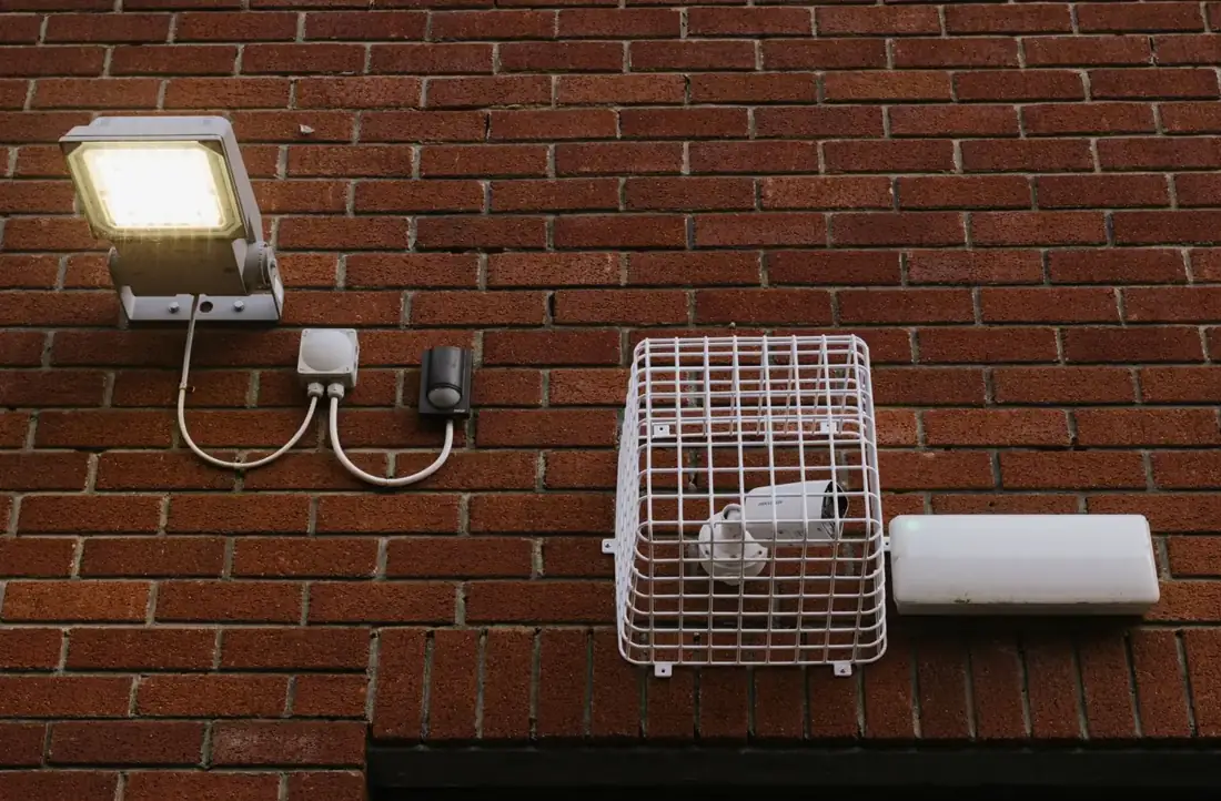LED floodlight with motion sensor beside a caged security camera on a brick wall